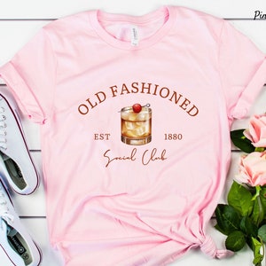 Puede incluir: Camiseta rosa con el texto "Old Fashioned Est 1880 Social Club" en letras marrones. Un dibujo de un c&oacute;ctel old fashioned con una cereza est&aacute; en el centro del texto.