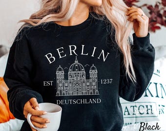 May include: Black sweatshirt with a white graphic design of the Berlin skyline, including the Brandenburg Gate, and the text "BERLIN EST 1237 DEUTSCHLAND".