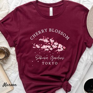 Puede incluir: Camiseta granate con el texto blanco "Cherry Blossom Sakura Gardens Tokyo" y un gr&aacute;fico de flores de cerezo rosas. La camiseta es de cuello redondo y tiene mangas cortas.