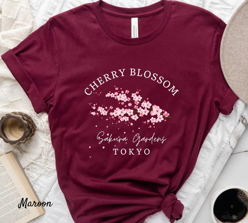 Puede incluir: Camiseta granate con el texto blanco "Cherry Blossom Sakura Gardens Tokyo" y un gr&aacute;fico de flores de cerezo rosas. La camiseta es de cuello redondo y tiene mangas cortas.
