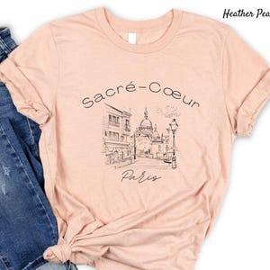 K&ouml;nnte beinhalten: Hellpfirsichfarbenes T-Shirt mit einer Schwarz-Wei&szlig;-Skizze der Basilika Sacr&eacute;-C&oelig;ur in Paris. Der Text "Sacr&eacute;-C&oelig;ur Paris" ist &uuml;ber der Illustration. Das T-Shirt hat kurze &Auml;rmel und einen Rundhalsausschnitt.