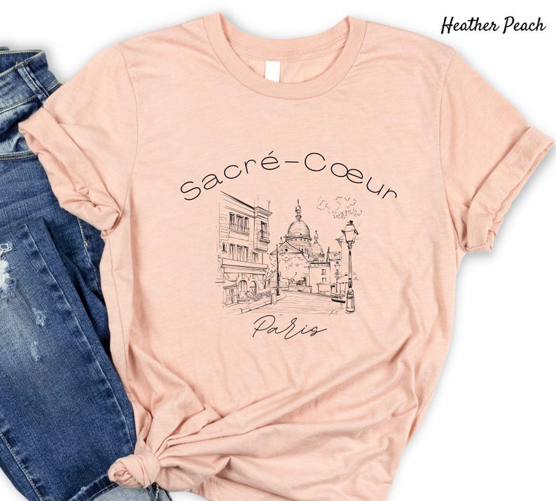 K&ouml;nnte beinhalten: Hellpfirsichfarbenes T-Shirt mit einer Schwarz-Wei&szlig;-Skizze der Basilika Sacr&eacute;-C&oelig;ur in Paris. Der Text "Sacr&eacute;-C&oelig;ur Paris" ist &uuml;ber der Illustration. Das T-Shirt hat kurze &Auml;rmel und einen Rundhalsausschnitt.