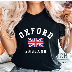 Puede incluir: Camiseta negra con texto blanco que dice "Oxford England" y una peque&ntilde;a bandera del Reino Unido.
