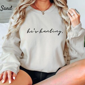 May include: Sand-colored sweatshirt with the text "he's hunting" in black script. The long-sleeved top is a neutral color and is suitable for casual wear. The word "Sand" is in the top left corner.