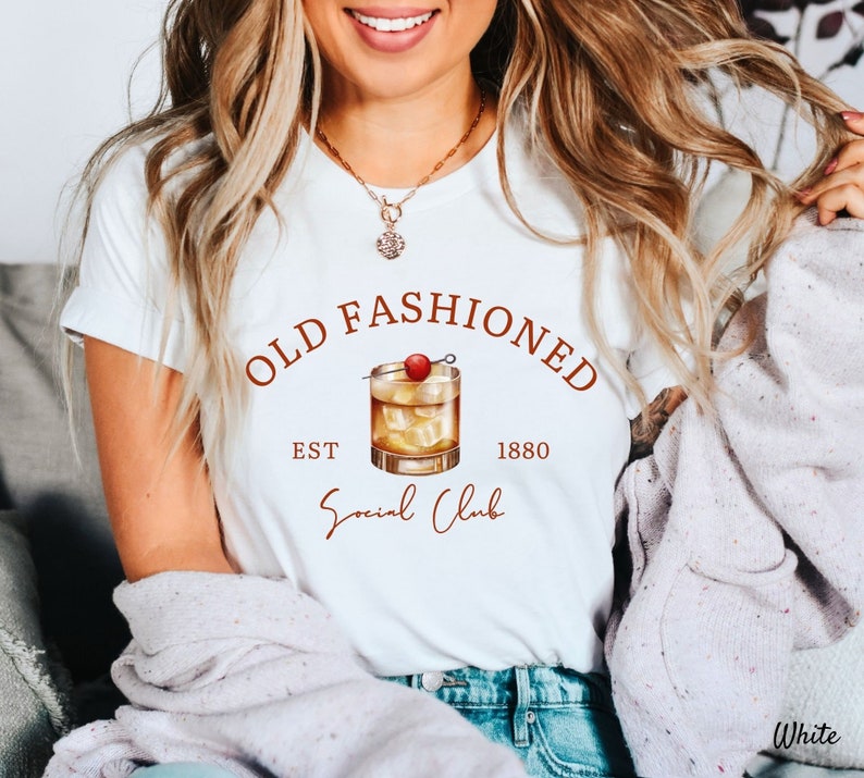 Puede incluir: Camiseta blanca con un gr&aacute;fico marr&oacute;n de un c&oacute;ctel old fashioned con una cereza y el texto "Old Fashioned Est 1880 Social Club".