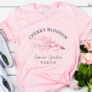 Puede incluir: Camiseta rosa claro con un dise&ntilde;o de flor de cerezo y la inscripci&oacute;n "Cherry Blossom Sakura Gardens Tokyo" en negro. Se ven zapatillas blancas.