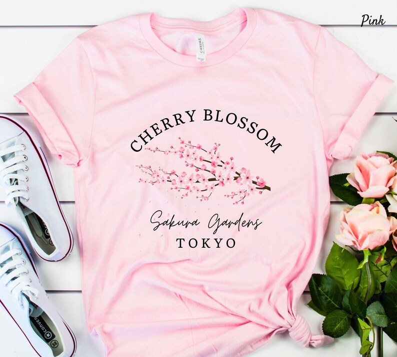 Puede incluir: Camiseta rosa claro con un dise&ntilde;o de flor de cerezo y la inscripci&oacute;n "Cherry Blossom Sakura Gardens Tokyo" en negro. Se ven zapatillas blancas.