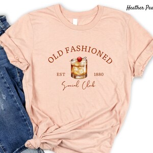Puede incluir: Una camiseta de color melocot&oacute;n p&aacute;lido con el texto "Old Fashioned Est 1880 Social Club" en letras marrones. Un dibujo de un c&oacute;ctel old fashioned con una cereza como adorno se encuentra en el centro del texto.
