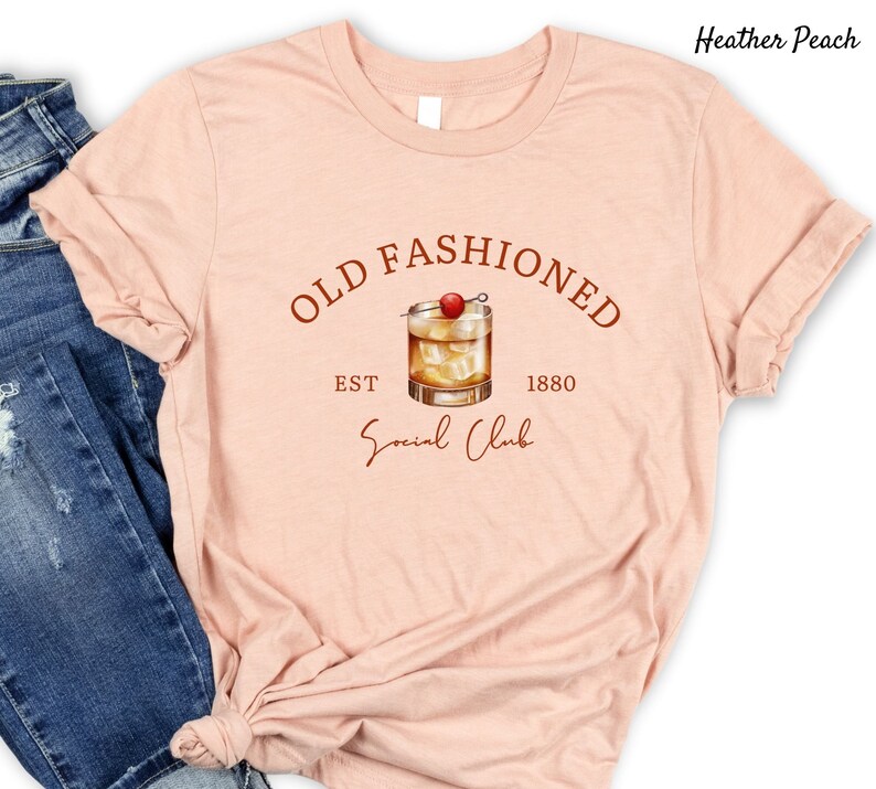 Puede incluir: Una camiseta de color melocot&oacute;n p&aacute;lido con el texto "Old Fashioned Est 1880 Social Club" en letras marrones. Un dibujo de un c&oacute;ctel old fashioned con una cereza como adorno se encuentra en el centro del texto.