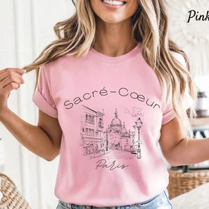 K&ouml;nnte beinhalten: Hellrosa T-Shirt mit einer schwarz-wei&szlig;en Illustration von Sacr&eacute;-C&oelig;ur in Paris. Die Worte "Sacr&eacute;-C&oelig;ur" und "Paris" sind auf dem Shirt aufgedruckt. Das T-Shirt hat einen Rundhalsausschnitt und hochgekrempelte &Auml;rmel.