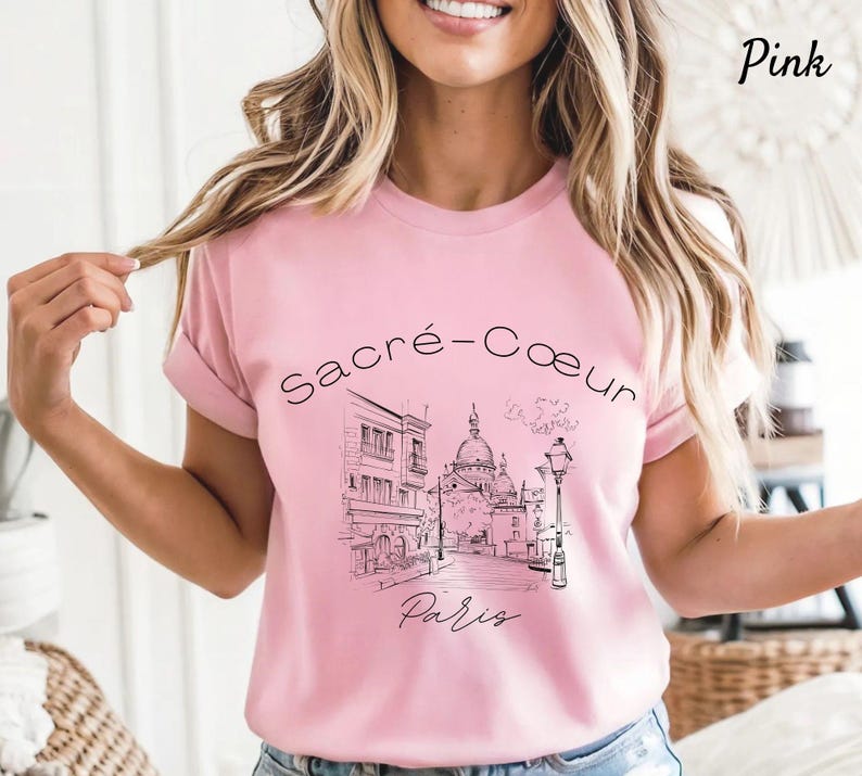 K&ouml;nnte beinhalten: Hellrosa T-Shirt mit einer schwarz-wei&szlig;en Illustration von Sacr&eacute;-C&oelig;ur in Paris. Die Worte "Sacr&eacute;-C&oelig;ur" und "Paris" sind auf dem Shirt aufgedruckt. Das T-Shirt hat einen Rundhalsausschnitt und hochgekrempelte &Auml;rmel.
