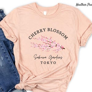 Puede incluir: Camiseta color melocot&oacute;n con dise&ntilde;o de flor de cerezo. El dise&ntilde;o incluye las palabras "Cherry Blossom" en semic&iacute;rculo sobre una rama con flores rosas. Debajo de la rama, las palabras "Sakura Gardens Tokyo" est&aacute;n escritas en cursiva.