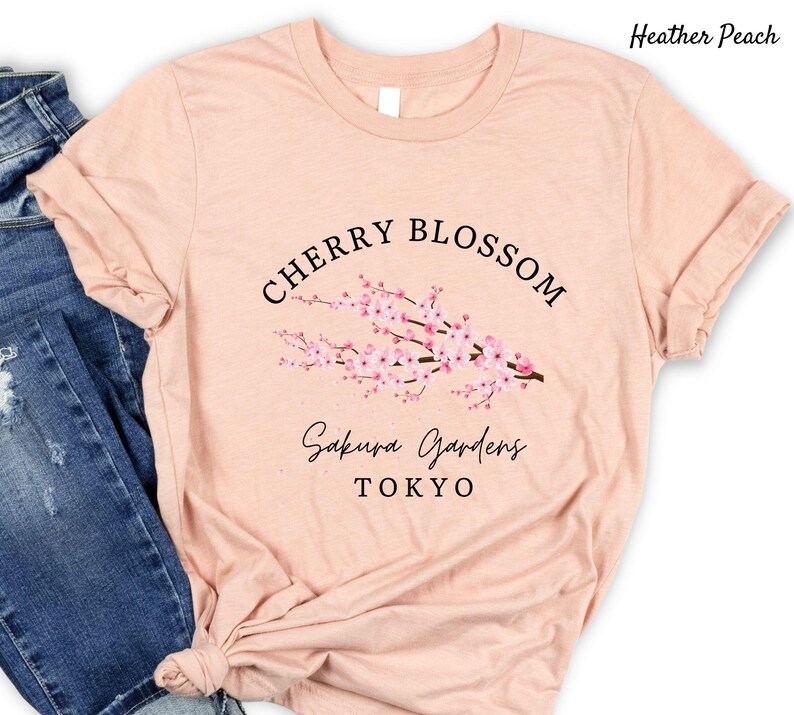 Puede incluir: Camiseta color melocot&oacute;n con dise&ntilde;o de flor de cerezo. El dise&ntilde;o incluye las palabras "Cherry Blossom" en semic&iacute;rculo sobre una rama con flores rosas. Debajo de la rama, las palabras "Sakura Gardens Tokyo" est&aacute;n escritas en cursiva.