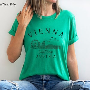 May include: A green t-shirt featuring a black graphic design of the Vienna skyline, including the Ferris wheel, St. Stephen's Cathedral, and the Hofburg Palace. The text "VIENNA", "Central Europe", and "AUSTRIA" is also included in the design.