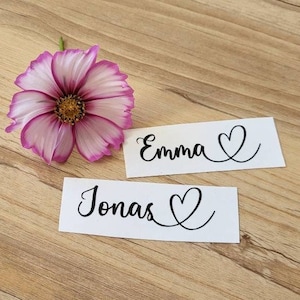 Könnte beinhalten: Zwei weiße Karten mit schwarzer Beschriftung. Die obere Karte sagt "Emma" mit einem Herzsymbol. Die untere Karte sagt "Jonas" mit einem Herzsymbol. Eine rosa Blume ist im Hintergrund.