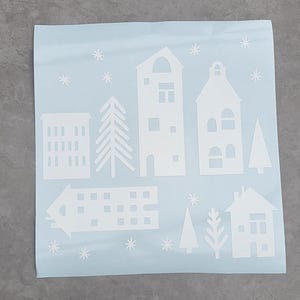Puede incluir: Una hoja de color azul claro con calcoman&iacute;as de vinilo blanco de varias formas de edificios arquitect&oacute;nicos, &aacute;rboles de hoja perenne y peque&ntilde;os patrones de copos de nieve en forma de estrella dispuestos sobre la superficie adhesiva cuadrada.