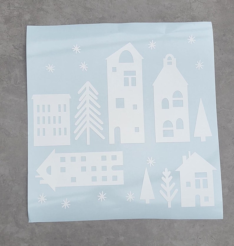 Puede incluir: Una hoja de color azul claro con calcoman&iacute;as de vinilo blanco de varias formas de edificios arquitect&oacute;nicos, &aacute;rboles de hoja perenne y peque&ntilde;os patrones de copos de nieve en forma de estrella dispuestos sobre la superficie adhesiva cuadrada.