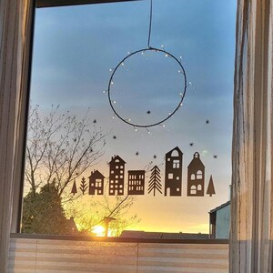 Puede incluir: Un adhesivo para ventana con una silueta de color marr&oacute;n oscuro de una hilera de casas, edificios y pinos, acentuada por peque&ntilde;as formas de estrellas. Sobre la escena del pueblo cuelga un marco de alambre circular adornado con luces blancas.