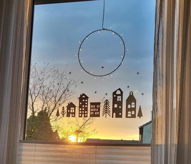 Puede incluir: Un adhesivo para ventana con una silueta de color marr&oacute;n oscuro de una hilera de casas, edificios y pinos, acentuada por peque&ntilde;as formas de estrellas. Sobre la escena del pueblo cuelga un marco de alambre circular adornado con luces blancas.