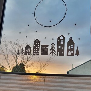 Puede incluir: Un adhesivo de ventana de color marr&oacute;n oscuro con la silueta de un peque&ntilde;o pueblo con varias formas de casas, pinos e iconos de estrellas dispersos. Sobre el pueblo, un fino marco circular de alambre cuelga de un hilo en el cristal.