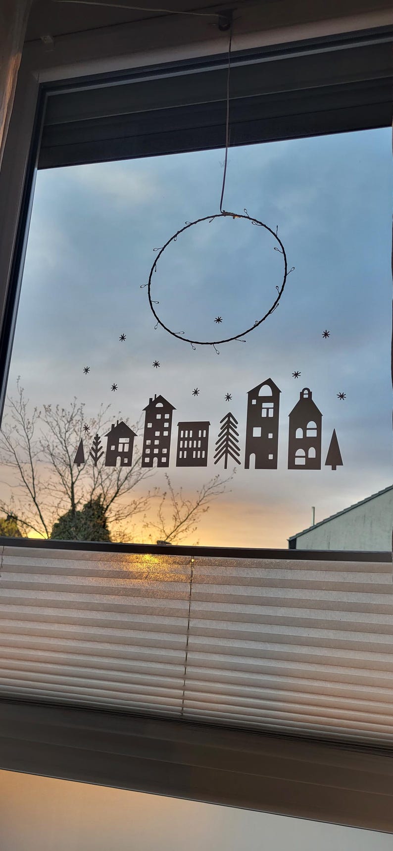 Puede incluir: Un adhesivo de ventana de color marr&oacute;n oscuro con la silueta de un peque&ntilde;o pueblo con varias formas de casas, pinos e iconos de estrellas dispersos. Sobre el pueblo, un fino marco circular de alambre cuelga de un hilo en el cristal.