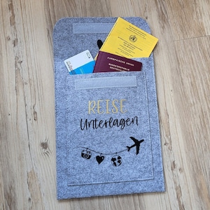 Könnte beinhalten: Grauer Filz-Reiseunterlagenhalter mit dem Text "Reise Unterlagen" in goldener Schrift. Der Halter hat ein Fach für Pässe, Tickets und andere Reiseunterlagen. Der Halter ist mit einem schwarzen Flugzeug, einer Kamera, einem Herzen und einem Globus verziert.