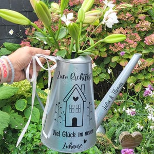 Könnte beinhalten: Eine silberne Gießkanne mit einem Hausdesign und dem Text "Zum Richtfest Viel Glück im neuen Zuhause" und "Julia & Thomas". Die Gießkanne ist mit weißen Blumen und einem weißen Band gefüllt.