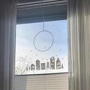 Puede incluir: Un adhesivo para ventana con la silueta de un peque&ntilde;o pueblo con casas, &aacute;rboles y estrellas dispersas, colocado debajo de un marco de alambre circular delgado que cuelga de la parte superior del marco de la ventana con un solo alambre.