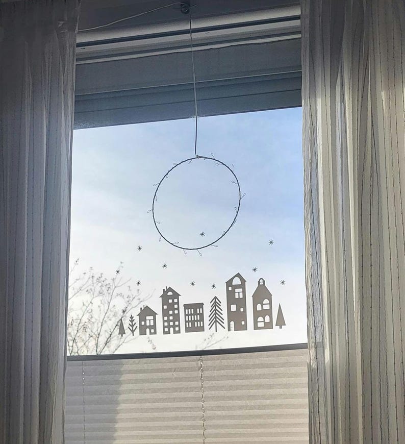Puede incluir: Un adhesivo para ventana con la silueta de un peque&ntilde;o pueblo con casas, &aacute;rboles y estrellas dispersas, colocado debajo de un marco de alambre circular delgado que cuelga de la parte superior del marco de la ventana con un solo alambre.