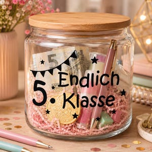 Könnte beinhalten: Ein durchsichtiges Glas mit Bambusdeckel, gefüllt mit rosa Papierstreifen, einem Fünf-Euro-Schein, einer Goldmünze und einem Stift. Auf der Vorderseite steht in schwarzer Schrift 5. Endlich Klasse, verziert mit kleinen Sternen und einer Wimpelkette.