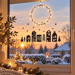 Puede incluir: Un adhesivo negro para ventana que muestra la silueta de un peque&ntilde;o pueblo con casas, &aacute;rboles y copos de nieve dispersos. Sobre el pueblo, una corona circular est&aacute; iluminada con luces de hadas de color blanco c&aacute;lido.