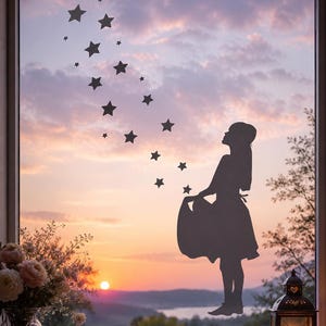 Könnte beinhalten: Ein dunkelgrauer Fensteraufkleber mit der Silhouette einer Person in einem Kleid, die den Saum des Rocks hält, mit einer Spur aus unterschiedlich großen Sternen, die sich von den Händen nach oben zur oberen linken Ecke erstreckt.