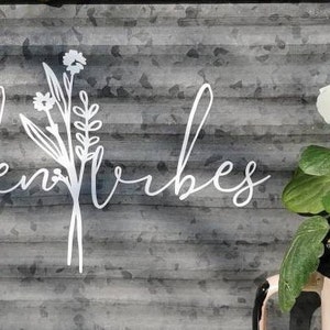 May include: A metal sign with the words "garden vibes" in cursive script, and a floral design. A potted pansy with white, purple, and yellow petals sits in a pink container, adding a touch of colour to the scene.