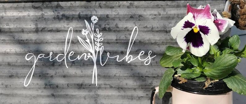 May include: A metal sign with the words "garden vibes" in cursive script, and a floral design. A potted pansy with white, purple, and yellow petals sits in a pink container, adding a touch of colour to the scene.