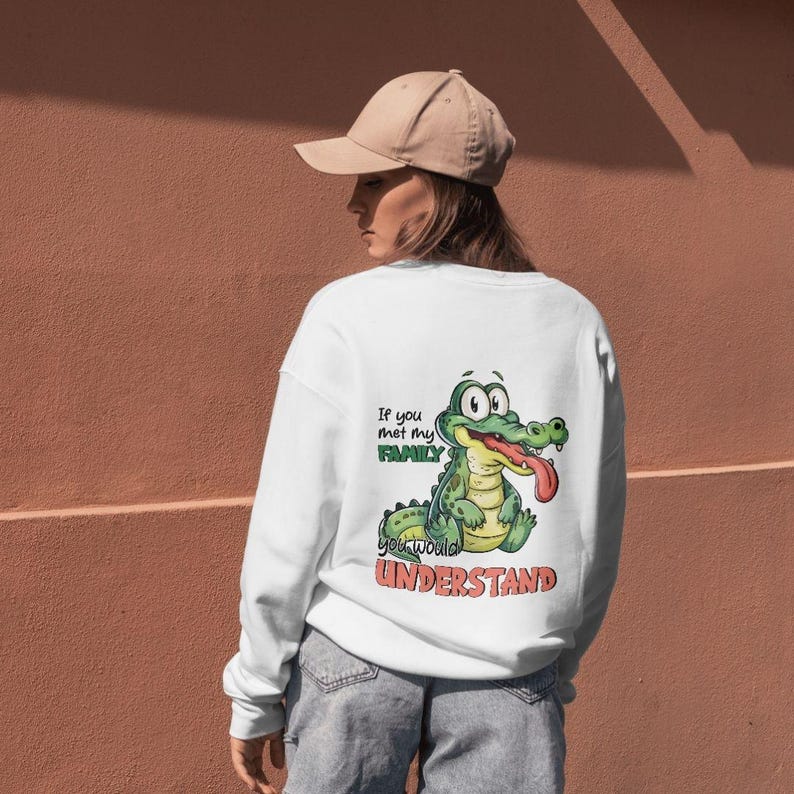 Puede incluir: Sudadera blanca con un dise&ntilde;o de cocodrilo de dibujos animados y el texto "If you met my FAMILY you would UNDERSTAND". El cocodrilo es verde con la lengua roja. La persona lleva una gorra beige.
