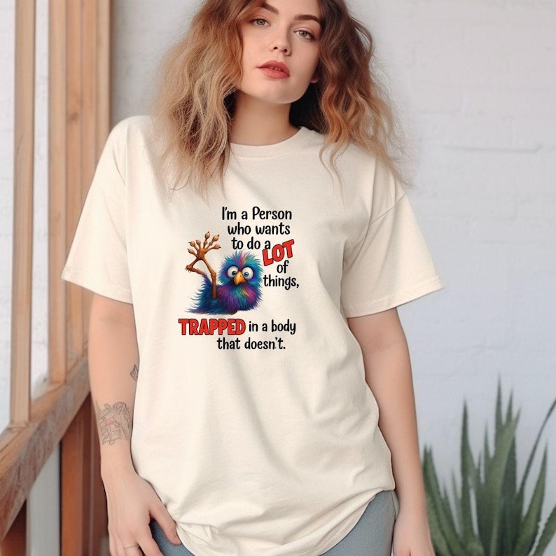 May include: Cream-colored t-shirt with a colorful cartoon creature graphic and the text "I'm a Person who wants to do a LOT of things, TRAPPED in a body that doesn't." The graphic features a blue, purple, and green creature.