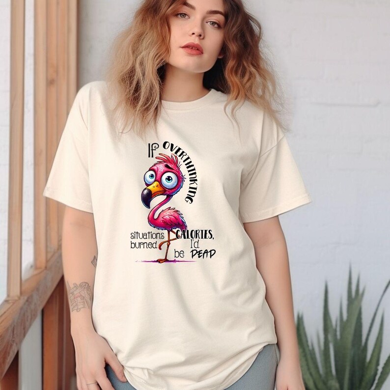 Puede incluir: Camiseta color crema con un flamenco de dibujos animados y el texto: "Si pensar demasiado en las situaciones quemara calor&iacute;as, estar&iacute;a muerto". El flamenco es rosa con pico amarillo y ojos azules. Estilo casual unisex.