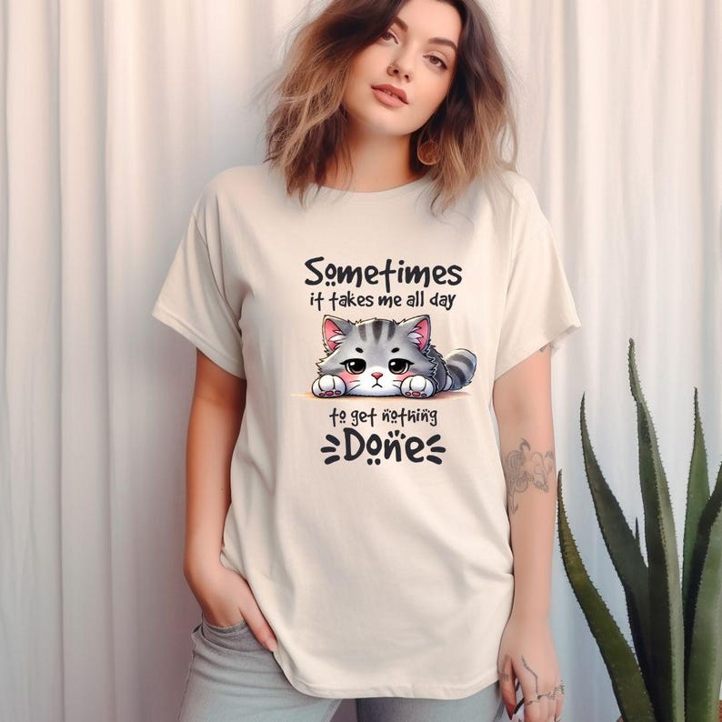 Puede incluir: Camiseta beige con una ilustraci&oacute;n de gato de dibujos animados y el texto "Sometimes it takes me all day to get nothing done." El gato es gris y blanco con acentos rosas. La camiseta tiene mangas cortas y un corte holgado.