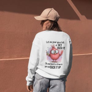 May include: White sweatshirt with a humorous graphic of a pink bird holding a drink, with the text "Let me pour you a tall of GET OVER IT. Oh and here's a straw so you can SUCK IT UP."