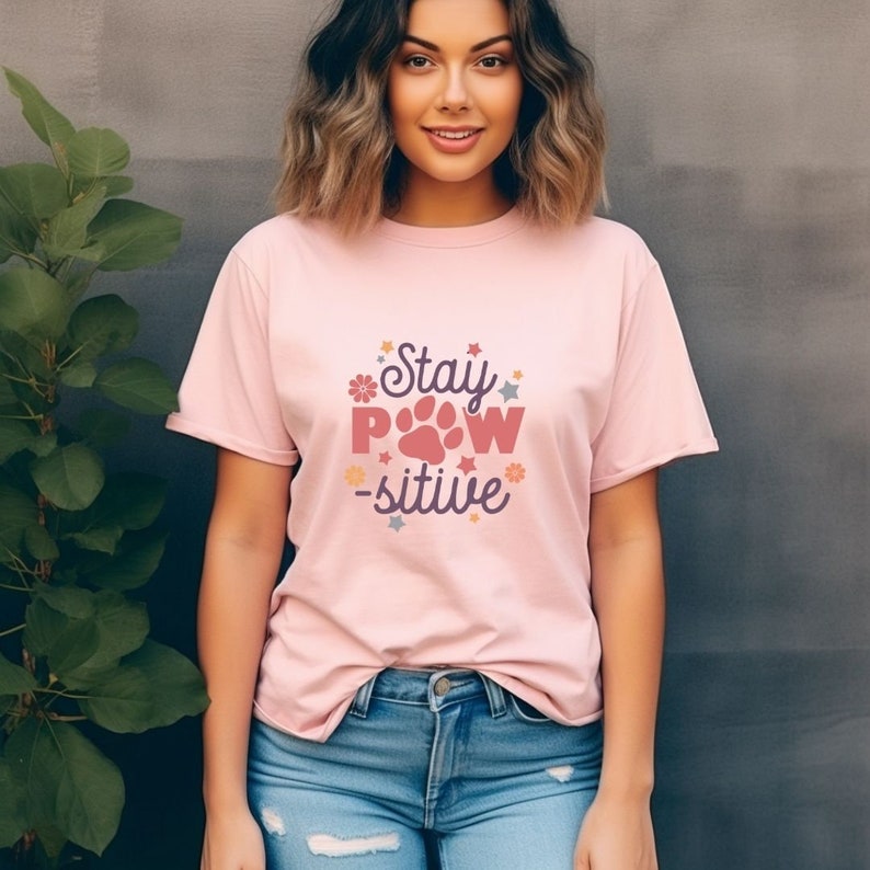 May include: A pink t-shirt with a graphic design that says "Stay Paw-sitive" with a paw print and flowers.