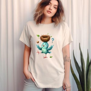 May include: Cream-colored t-shirt featuring a cartoon bird with a nest on its head. The bird is blue with outstretched wings and a confused expression. A brown nest, green leaves, and a question mark are also visible.