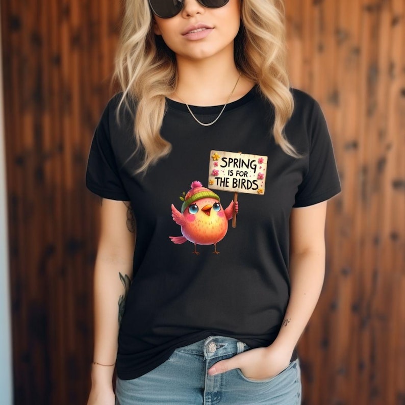 May include: Black t-shirt featuring a colorful cartoon bird holding a sign that reads "SPRING IS FOR THE BIRDS." The bird is wearing a green hat and the sign has floral accents. The shirt is a classic crew neck.