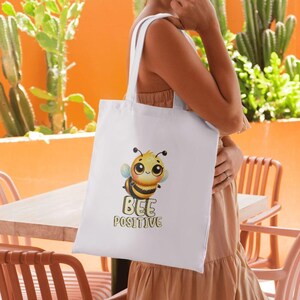 Puede incluir: Bolso tote blanco con una ilustraci&oacute;n de abeja de dibujos animados. La abeja tiene un cuerpo a rayas amarillas y negras y ojos grandes. El texto "BEE POSITIVE" est&aacute; debajo. El bolso tiene dos asas blancas.
