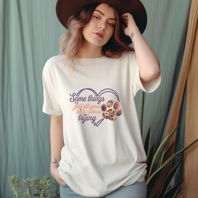 May include: White t-shirt with a purple heart design featuring a paw print with floral accents. The text "Some things just fill your heart without trying" is printed on the shirt.