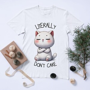 Puede incluir: Camiseta blanca con un gato de dibujos animados y el texto "LITERALLY DON'T CARE". El gato tiene los ojos cerrados, los brazos cruzados y detalles en rosa. La camiseta est&aacute; sobre una superficie blanca con elementos decorativos.