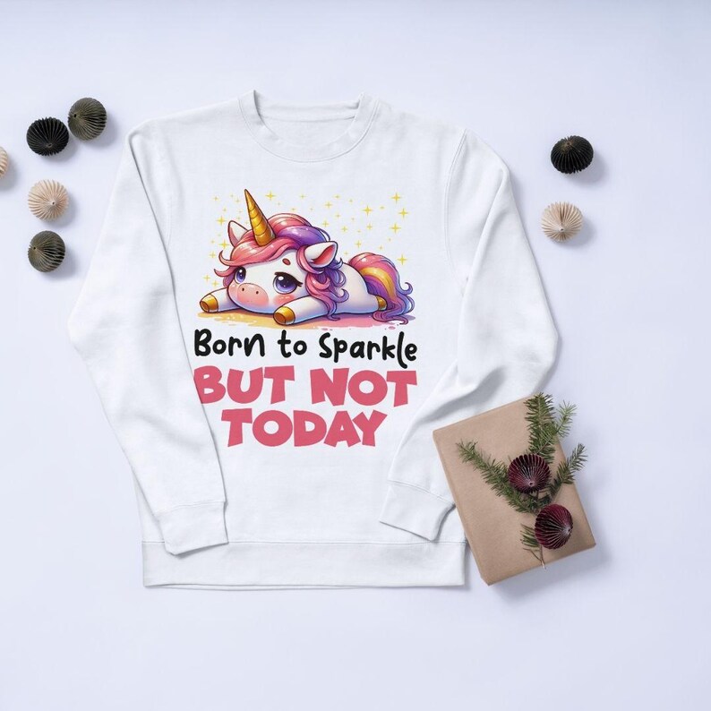 Puede incluir: Sudadera blanca con un dise&ntilde;o de unicornio de dibujos animados, con una melena rosa y morada y un cuerno dorado. El texto "Born to Sparkle BUT NOT TODAY" est&aacute; impreso debajo. Una prenda de vestir casual.