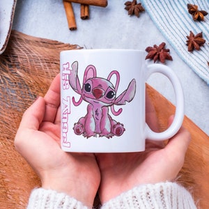 以下が含まれることがあります： ピンクのスティッチのキャラクターイラストとピンクの文字で「His Angel」と書かれた白いセラミックマグカップです。