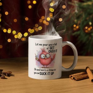 May include: White ceramic mug with a humorous cartoon bird design. The bird is pink with large eyes and is holding a drink. Text on the mug reads "Let me pour you a tall of GET OVER IT" and "Oh and here's a straw so you can SUCK IT UP."