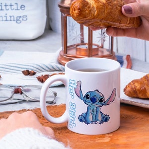 以下が含まれることがあります： リロ＆スティッチの青いスティッチのキャラクターが描かれた白いセラミックマグカップ。マグカップには「Her Stitch」の文字が側面に書かれています。