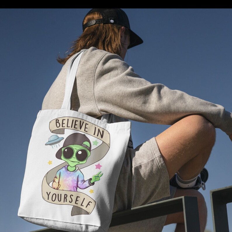 May include: A white tote bag featuring a green alien cartoon character wearing a tie-dye shirt and a backwards baseball cap. The alien is holding up a peace sign. The bag has the text "Believe in Yourself" on it.